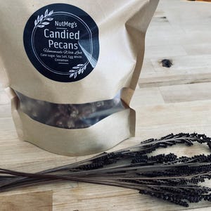 Könnte beinhalten: Eine braune Papiertüte mit kandierten Pekannüssen und einem transparenten Fenster. Das Etikett in Schwarz und Weiß lautet "NutMeg's Candied Pecans". Zutaten: Rohrzucker, Meersalz, Eiweiß und Zimt. Getrocknete Stiele liegen im Vordergrund.