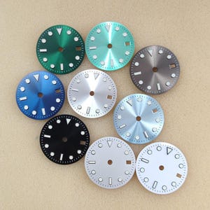 Puede incluir: Una colección de nueve esferas de reloj circulares en varios colores, incluyendo verde, azul claro, azul oscuro, negro, plata, blanco y gris. Cada esfera presenta un orificio central, una ventana de fecha e índices blancos con un triángulo en las doce.