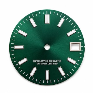 May include: A circular green watch dial with a sunburst finish, featuring silver-tone applied hour markers with white luminous inserts, a date window at the three o'clock position, and the text SUPERLATIVE CHRONOMETER OFFICIALLY CERTIFIED.