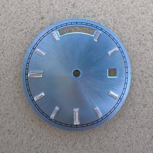 Pode incluir: Um mostrador de relógio circular azul-claro com acabamento em metal escovado. Apresenta marcadores de hora retangulares em tom prateado, um orifício central para os ponteiros, uma janela de data retangular às três horas e um minutero preto.
