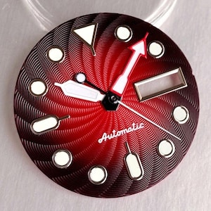 Puede incluir: Una esfera de reloj circular con un degradado de rojo intenso a negro y un patrón texturizado en espiral. Incluye marcadores de hora luminiscentes blancos, una ventana de fecha rectangular y la palabra Automatic en letra blanca.