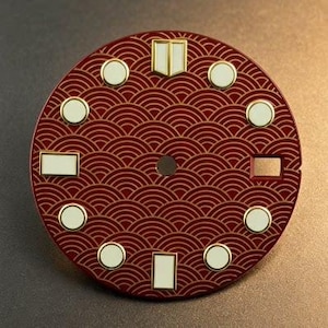 Pode incluir: Um mostrador de relógio circular com fundo vermelho profundo e um padrão repetitivo de ondas douradas. O mostrador inclui marcadores de hora circulares brancos, marcadores retangulares às doze, seis e nove horas, e uma janela de data às três.