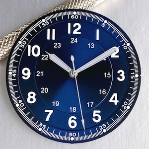 Peut inclure: Un cadran de montre circulaire avec une finition bleu profond soleillée, affichant des chiffres arabes blancs de 1 à 12 et une échelle intérieure de 24 heures de 13 à 24. Le cadran présente des aiguilles blanches et une graduation des minutes.