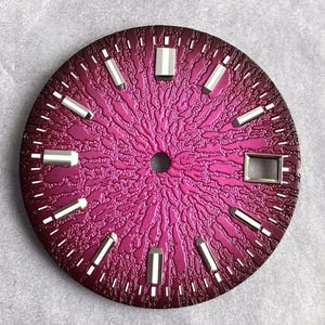 Pode incluir: Um mostrador de relógio circular com um gradiente vibrante de magenta e roxo profundo com um padrão radial orgânico texturizado. Inclui marcadores de hora retangulares prateados e uma moldura de janela de data retangular às três horas.