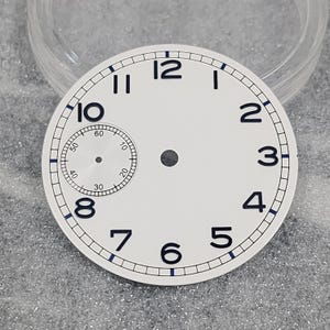 Puede incluir: Una esfera de reloj redonda y blanca con números arábigos negros del 2 al 12 y el 8. Una subesfera más pequeña está situada en la posición de las 9, mostrando los números 10, 20, 30, 40, 50 y 60. El borde exterior tiene una pista de minutos negra y azul.