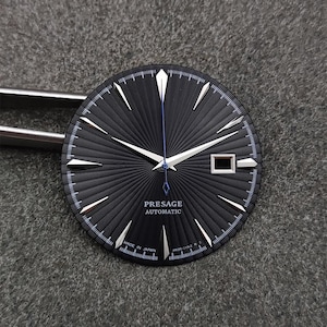 Puede incluir: Una esfera de reloj negra redonda con patrón de rayos de sol, marcadores de hora plateados y manecillas plateadas. Un segundero azul fino sale del centro. El texto dice PRESAGE AUTOMATIC, MADE IN JAPAN y 4R35-01N2 R 2. Una ventana de fecha está a las 3.