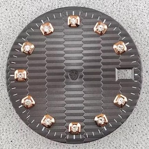 Peut inclure: Un cadran de montre circulaire gris foncé avec un motif horizontal texturé. Le cadran comprend dix index carrés en or rose sertis de pierres claires, des index des minutes imprimés en blanc et un guichet de date rectangulaire à trois heures.