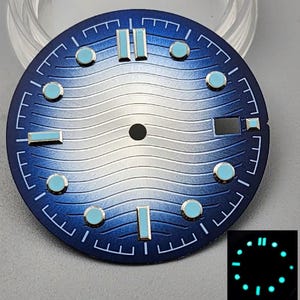 May include: A circular watch dial featuring a blue gradient finish with a wavy texture pattern. It includes light blue hour markers, a central hole for hands, and a rectangular date window at the three o'clock position. An inset shows the dial glowing.
