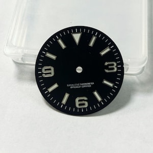 May include: A black watch face with white hour markers and numerals 3, 6, and 9. The text "SUPERLATIVE CHRONOMETER OFFICIALLY CERTIFIED" is printed at the bottom. The face is round and has a small hole in the center.