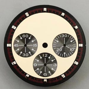 Puede incluir: Una esfera de reloj redonda con un centro color crema y tres subesferas gris oscuro. Las subesferas muestran los números 60, 45, 30, 15, 30, 20, 10, 12, 9, 6 y 3. Un anillo exterior negro contiene marcas rojas e índices plateados. El texto JAPAN MOVT está impreso en la parte inferior.