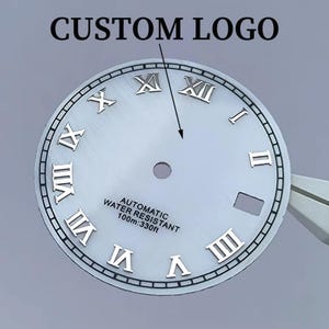 Può includere: Un quadrante circolare argentato con numeri romani in rilievo, un foro centrale e una finestrella per la data. Il testo recita AUTOMATIC WATER RESISTANT 100m:330ft. Una freccia punta verso l'alto con il testo CUSTOM LOGO.