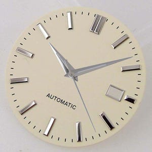 Puede incluir: Una esfera de reloj redonda con fondo color crema, marcadores de hora aplicados en tono plateado y manecillas plateadas. La palabra AUTOMATIC está impresa en negro debajo del centro. Una ventana de fecha rectangular se sitúa a las tres.