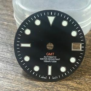 Op de afbeelding: Een zwarte wijzerplaat met witte uurmarkeringen en een datumvenster. De wijzerplaat heeft de tekst "GMT AUTOMATIC WATER RESISTANT 10BAR/100M" in rood en wit. De wijzerplaat heeft ook de tekst "JAPAN" en "MOVT".