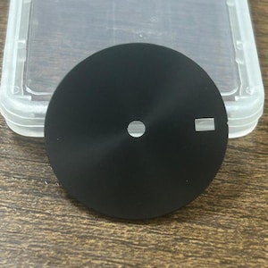 Könnte beinhalten: Ein schwarzes Zifferblatt mit einem kleinen Loch und einem rechteckigen Marker. Das Zifferblatt ist kreisförmig und hat eine matte Oberfläche. Es liegt neben einem transparenten Kunststoffbehälter.