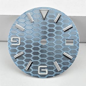 Peut inclure: Un cadran de montre circulaire arborant un motif en nid d'abeille bleu clair avec une finition métallique brossée. Il comprend des index appliqués argentés, un marqueur triangulaire à douze heures et les chiffres 6 et 9.