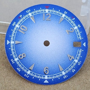 Pode incluir: Um mostrador de relógio circular azul com acabamento degradê, apresentando numerais aplicados em tom prateado às 12, 2, 4, 6, 8 e 10 horas. A borda externa exibe uma escala de bússola branca com direções cardeais e graus, além de uma data.