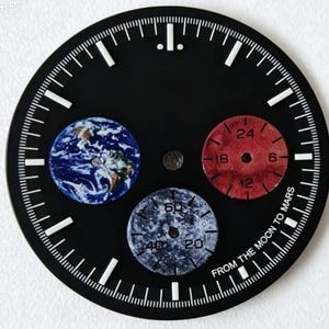 Peut inclure: Un cadran de montre noir rond avec des index blancs et trois sous-cadrans. Un sous-cadran affiche une image de la Terre bleue et blanche, un autre est une échelle rouge de 24 heures, et le troisième est une échelle grise texturée de 60 secondes.