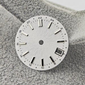 Peut inclure: Un cadran de montre blanc avec des index argentés et un centre texturé rayonnant vers l'extérieur. Le cadran comporte des chiffres noirs et un guichet de date à 6 heures. Le cadran est sur une surface grise.