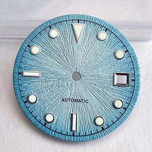 Puede incluir: Una esfera de reloj circular de color azul claro con un patrón texturizado de rayos de sol que irradia desde el centro. Incluye marcadores de hora luminiscentes blancos de varias formas, una ventana de fecha cuadrada y la palabra AUTOMATIC.