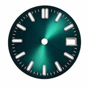 Può includere: Un quadrante per orologio circolare con finitura verde smeraldo a raggiera. Presenta indici applicati color argento con centri luminosi bianchi, un foro centrale per le lancette e una finestrella rettangolare per la data a ore tre.