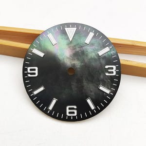 Puede incluir: Una esfera de reloj redonda con una superficie de nácar iridiscente oscura con degradado negro. Incluye marcadores de hora luminiscentes blancos, un índice triangular en la posición de las doce y números arábigos blancos en el tres, seis y nueve.