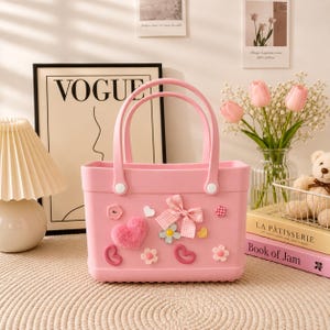 May include: A pink tote bag with two handles, decorated with a plush heart, a gingham bow, and floral and heart-shaped embellishments. The bag is sitting on a beige surface.