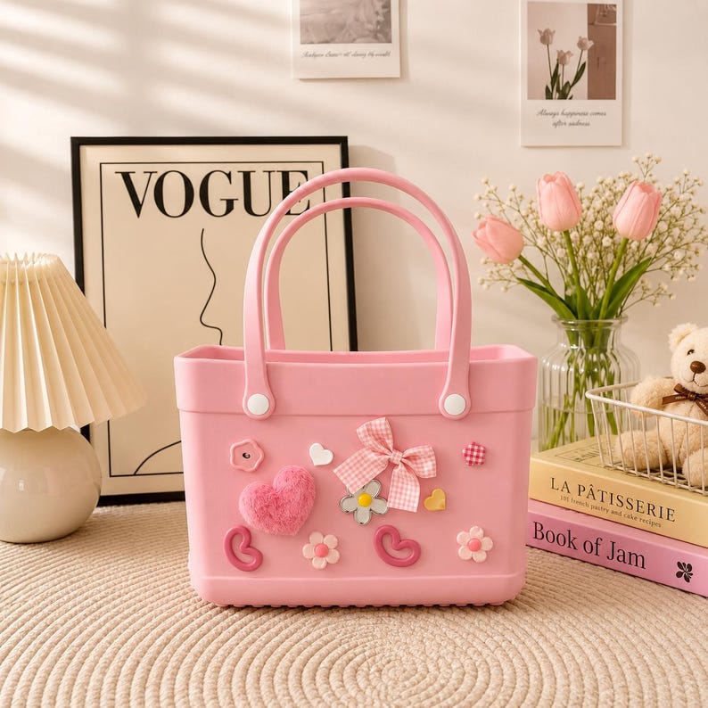 May include: A pink tote bag with two handles, decorated with a plush heart, a gingham bow, and floral and heart-shaped embellishments. The bag is sitting on a beige surface.