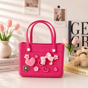 May include: A bright pink tote bag with two handles and decorative embellishments. The bag features a fluffy pink heart, a gingham bow, and various flower and heart-shaped accents. The bag is sitting on a white surface.