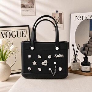 May include: Black tote bag with two handles and decorative embellishments. The bag features a variety of rhinestone accents, including a star, heart, and the word "Queen". The bag has a rectangular shape.