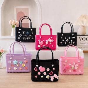 May include: A collection of colorful tote bags in black, pink, and lavender. Each bag features decorative embellishments like stars, hearts, bows, and buttons. The bags have matching handles and are displayed on a wooden surface.