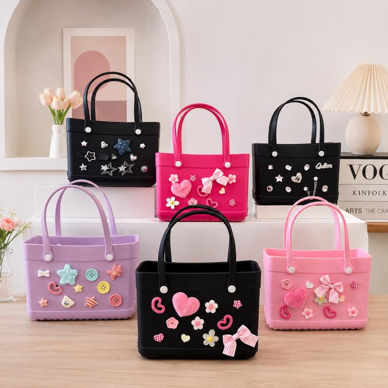 May include: A collection of colorful tote bags in black, pink, and lavender. Each bag features decorative embellishments like stars, hearts, bows, and buttons. The bags have matching handles and are displayed on a wooden surface.
