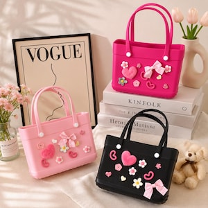 May include: Three small tote bags in pink and black, decorated with bows, hearts, and flowers. The bags have matching handles and are displayed with a Vogue print and books. A teddy bear and flowers are also in the image.