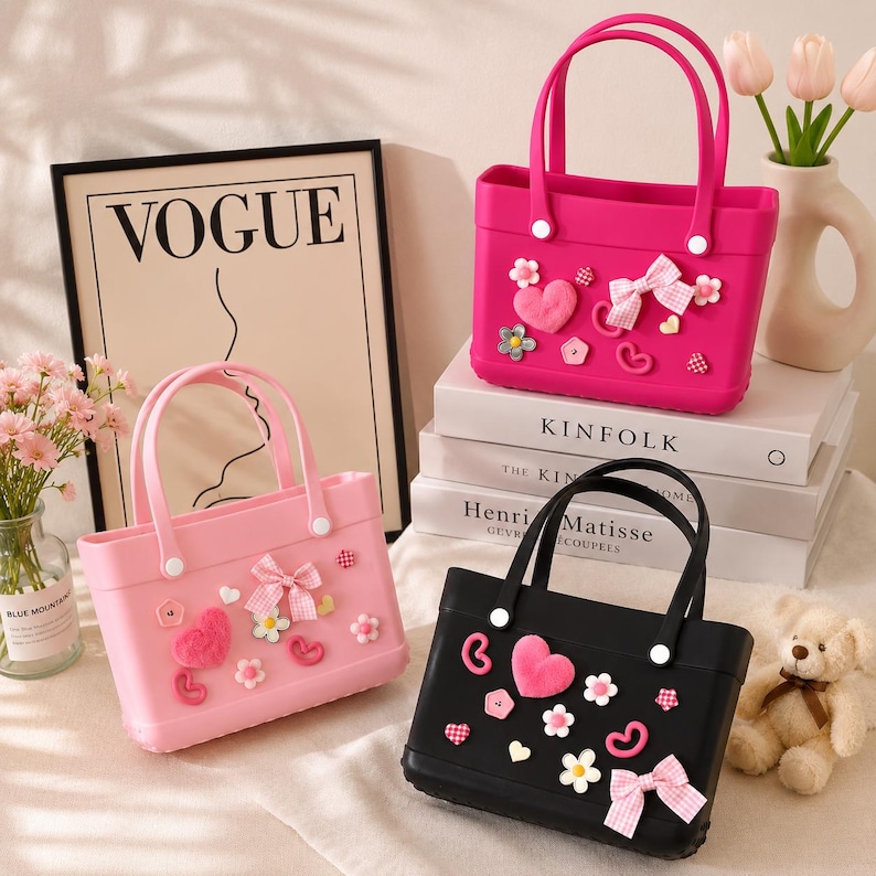 May include: Three small tote bags in pink and black, decorated with bows, hearts, and flowers. The bags have matching handles and are displayed with a Vogue print and books. A teddy bear and flowers are also in the image.