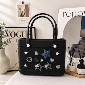 May include: Black tote bag with two handles and decorative embellishments. The bag features a variety of star, heart, and smiley face charms. The bag has a rectangular shape and white accents on the handles.