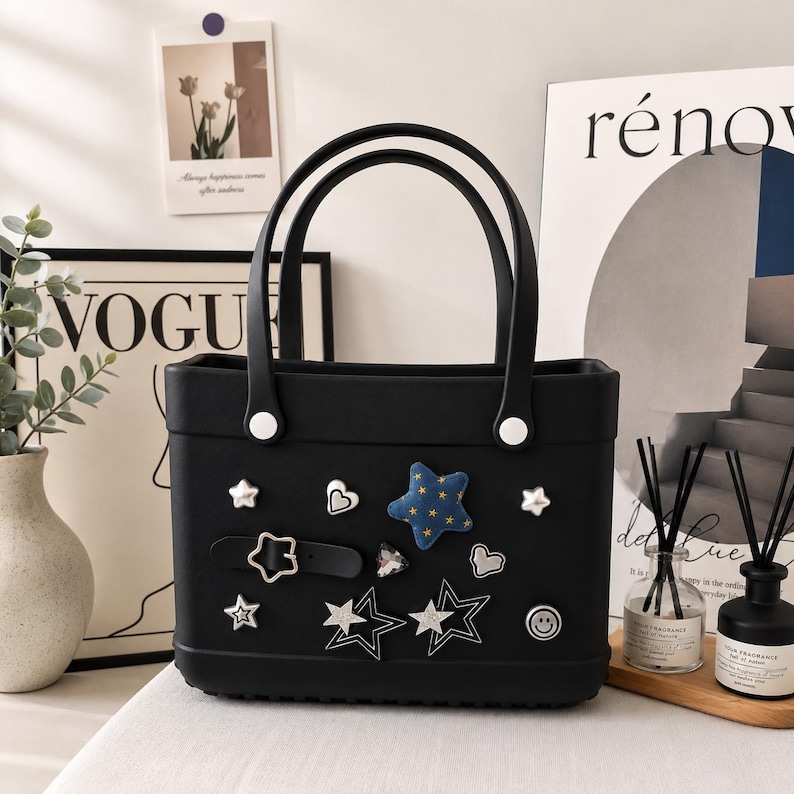 May include: Black tote bag with two handles and decorative embellishments. The bag features a variety of star, heart, and smiley face charms. The bag has a rectangular shape and white accents on the handles.
