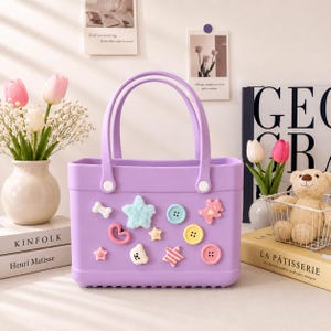 May include: A lavender tote bag with two handles and decorative embellishments. The bag features a bone, star, heart, and button-shaped decorations in various colors. The bag is sitting next to a vase of flowers and a book.