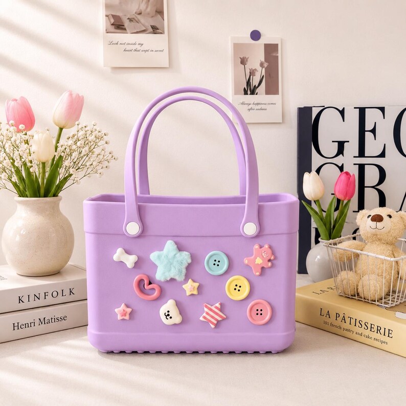 May include: A lavender tote bag with two handles and decorative embellishments. The bag features a bone, star, heart, and button-shaped decorations in various colors. The bag is sitting next to a vase of flowers and a book.