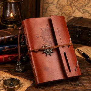 Op de afbeelding: Een bruin lederen dagboek met een in reliëf aangebracht kompasroosontwerp op de voorkant. Het dagboek wordt gesloten met een leren koord dat om een kleine metalen scheepsroerbedel is gewikkeld. Decoratieve metalen ankerbedels hangen aan de zijkant.