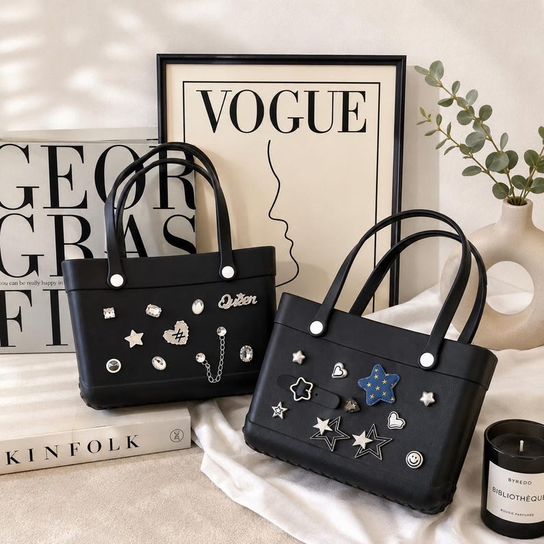 May include: Two black tote bags with white handles and decorative embellishments. One bag has a "Queen" charm and various rhinestone and chain accents. The other bag features star, heart, and smiley face charms. A "VOGUE" print is in the background.