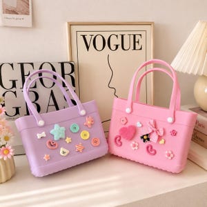 May include: Two small, rectangular tote bags. One is lavender, the other is pink. Both have matching handles and are decorated with various embellishments, including buttons, stars, and hearts. The bags are sitting on a white surface.