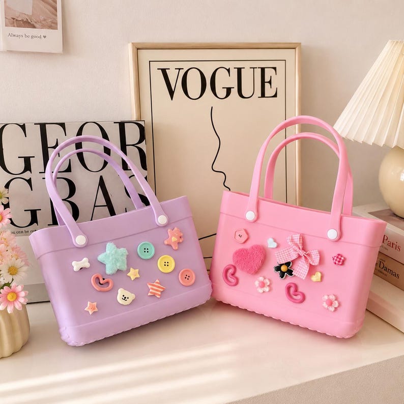 May include: Two small, rectangular tote bags. One is lavender, the other is pink. Both have matching handles and are decorated with various embellishments, including buttons, stars, and hearts. The bags are sitting on a white surface.