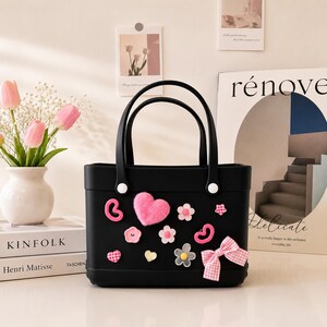 May include: Black tote bag with decorative pink heart, flower, and bow accents. The bag has two handles and white button details. A vase of pink tulips and books are in the background.