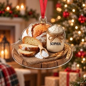 May include: A clear acrylic Christmas ornament featuring a printed image of a loaf of crusty bread, three bread slices, a glass jar filled with flour labeled SOPHIA, a small wooden scoop with flour, and wheat stalks on a burlap-covered wooden board.