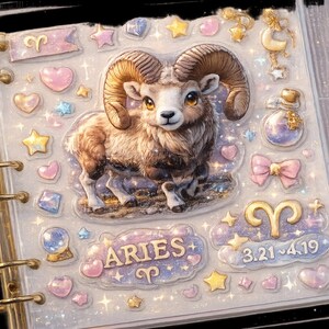 May include: A clear planner with gold rings, decorated with an Aries theme. The cover shows a detailed ram illustration, surrounded by stars, hearts, and the Aries symbol. The text "ARIES" and the dates 3.21-4.19 are also visible.
