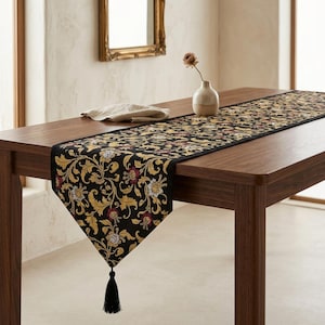 May include: A black table runner featuring a gold and burgundy floral scroll pattern with a pointed end finished with a black tassel. The runner is draped over a wooden table alongside a beige napkin and a small ceramic vase.