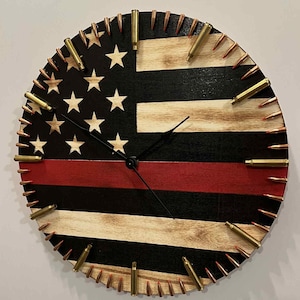 May include: Round wooden wall clock featuring an American flag design with a red stripe. The clock face has white stars on a black background and is accented with brass bullet casings around the perimeter. The clock hands are black.