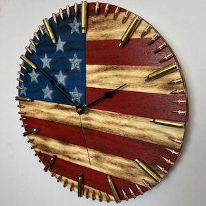May include: A round wooden clock with a painted American flag design. The clock face is divided into red and white stripes and a blue field with white stars. Brass bullet casings are embedded around the clock's edge, serving as hour markers.