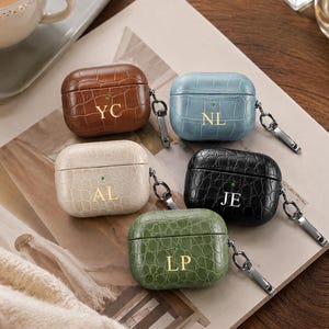 May include: Five rectangular earbud cases with a crocodile-embossed texture in brown, light blue, cream, black, and olive green. Each case features gold-toned initials, a small circular LED indicator light, and a metal carabiner clip attached to the side.