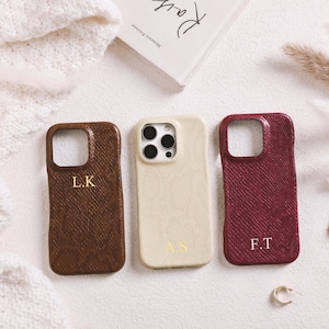 May include: Three personalized phone cases in brown, cream, and burgundy, each with a textured finish and gold initials. The cases are arranged on a white surface with a book and earrings.