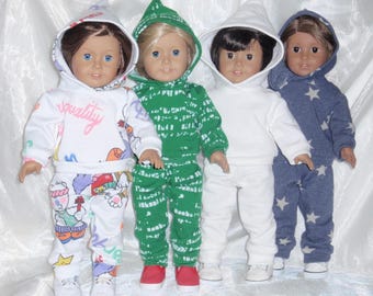 May include: Four dolls wearing hooded sweatshirts and pants. The dolls are wearing different colored outfits, including white with a pink and blue graphic, green with a black and white graphic, white with a white graphic, and blue with a white star graphic.